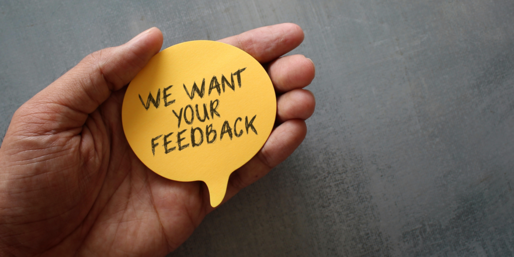 A hand holding a speech-bubble shaped post-it note saying "We Want Your Feedback"